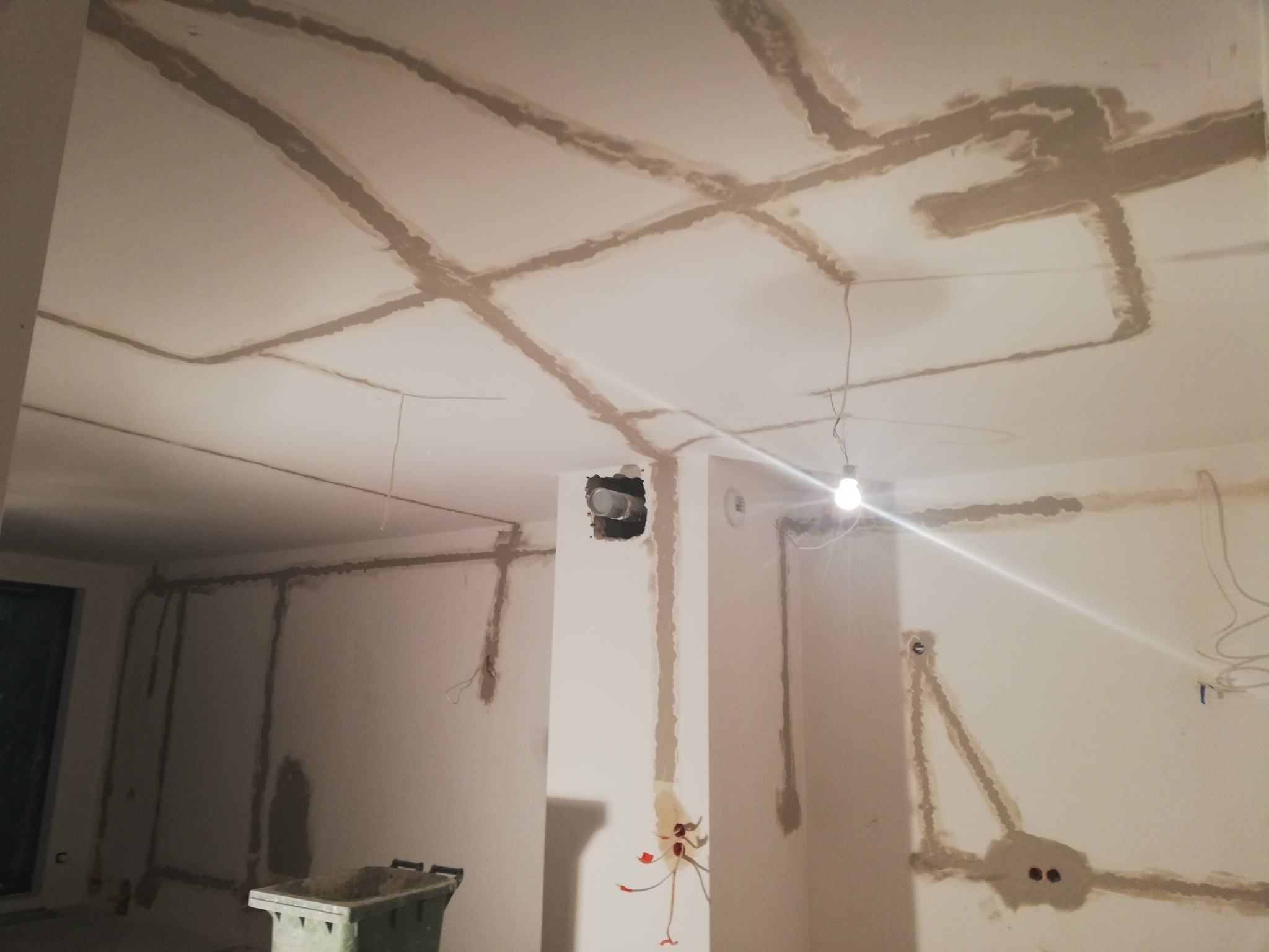 Przygotowanie ścian i sufitu do prac wykończeniowych, widoczne bruzdy wypełnione masą szpachlową pod instalacje elektryczne, surowy stan wnętrza z wiszącą żarówką.