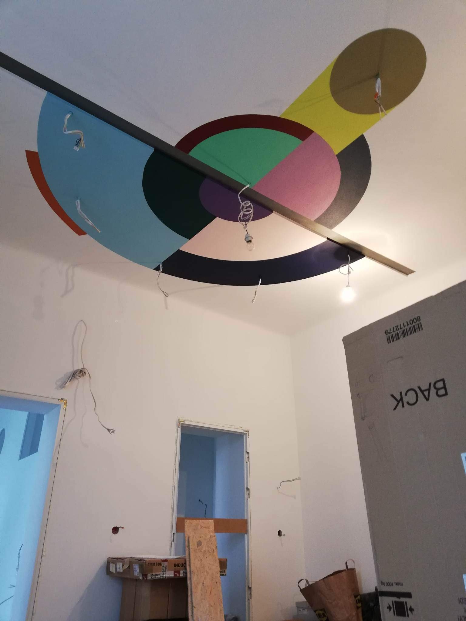 Niedokończony sufit z geometrycznym wzorem w kolorach pastelowych, przecięty drewnianą belką z wiszącymi żarówkami, widoczne kable i kartony w pomieszczeniu w trakcie prac wykończeniowych.