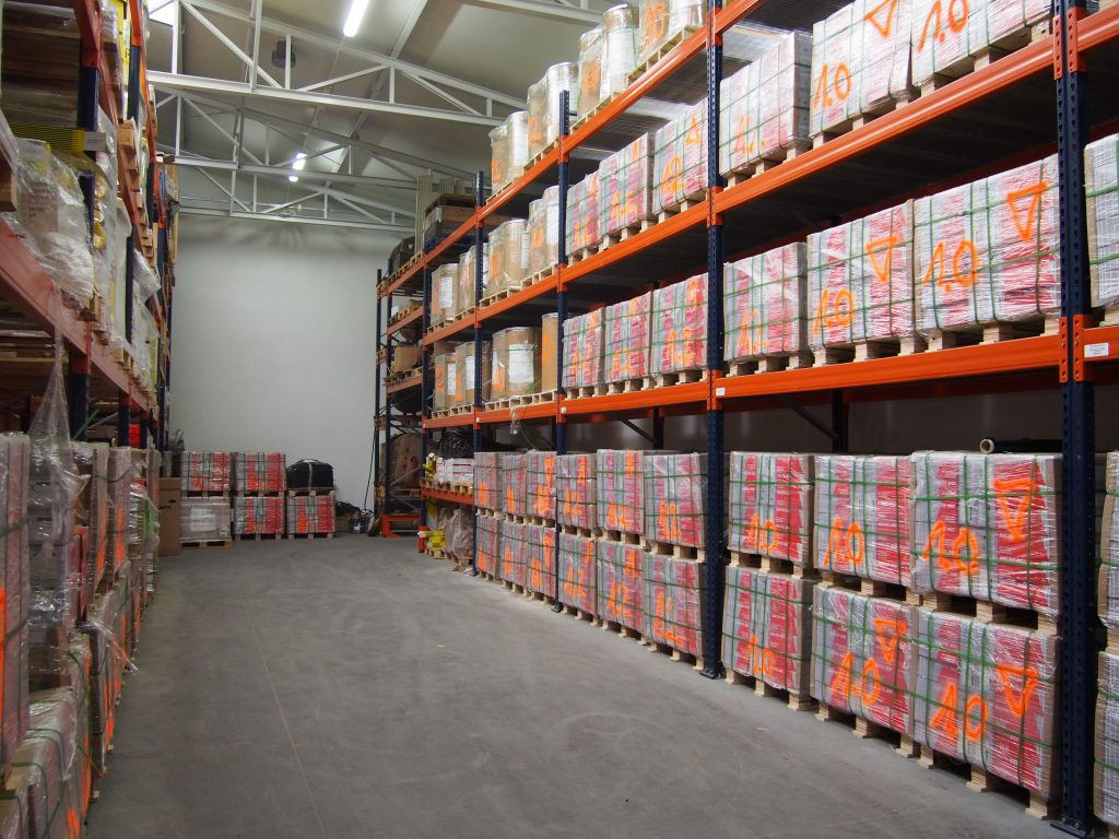 Pełny magazyn z paletami cegieł, każda owinięta folią i oznaczona pomarańczowym numerem '40', ustawionymi na metalowych regałach pod jasnym, przemysłowym oświetleniem.