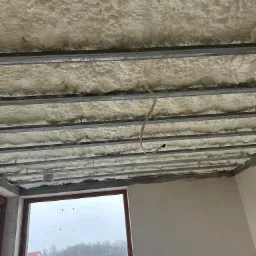 Szczegółowe ujęcie izolacji poddasza z pianki poliuretanowej, widoczne metalowe profile konstrukcyjne, kabel zwisający z sufitu, okno z widokiem na krajobraz za oknem