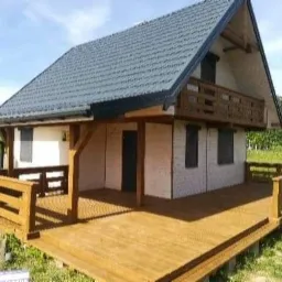 Gotowy, parterowy dom z bali z szarym dachem dwuspadowym, drewnianym tarasem i balkonem na piętrze, widok z zewnątrz w słoneczny dzień.