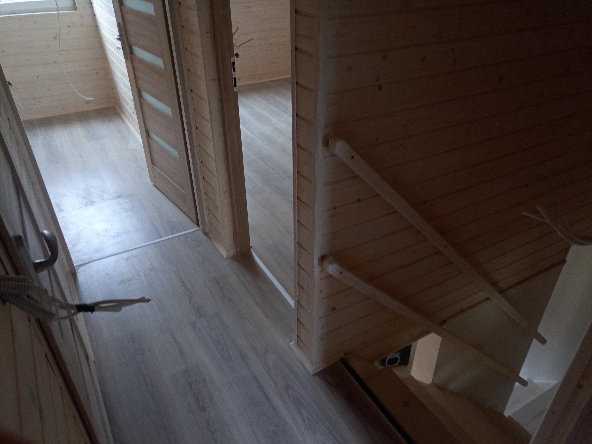 Wnętrze domu z drewnianych paneli, widok na korytarz z podłogą imitującą drewno i schody w dół, drzwi wewnętrzne z przeszkleniami.