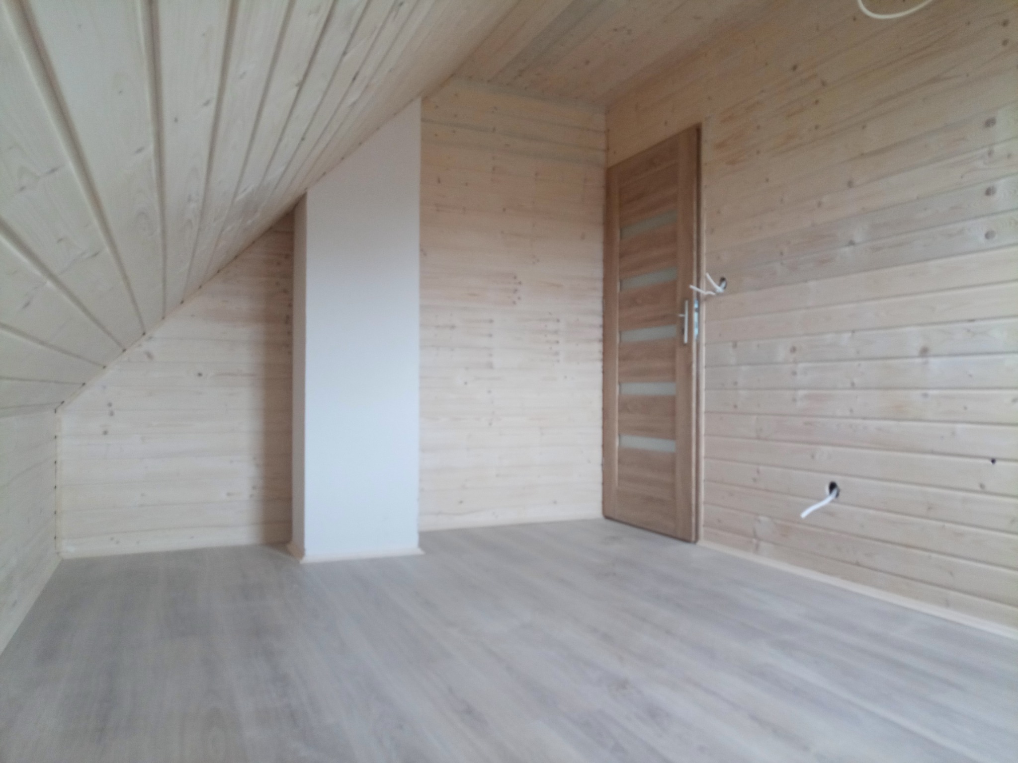 Pokój na poddaszu wykończony jasnym drewnem, z ukośnym sufitem, białą ścianką działową i drewnianą podłogą, widoczna również drewniana ościeżnica drzwiowa z częściowo zamontowanymi drzwiami.
