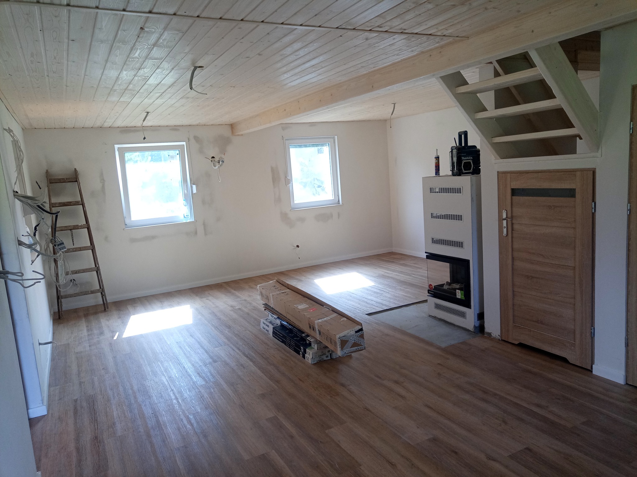 Wnętrze domu w trakcie wykańczania: ułożona podłoga z paneli imitujących drewno, częściowo wykończone ściany, drewniany strop, schody w surowym stanie, drabina oparta o ścianę, paczki paneli...