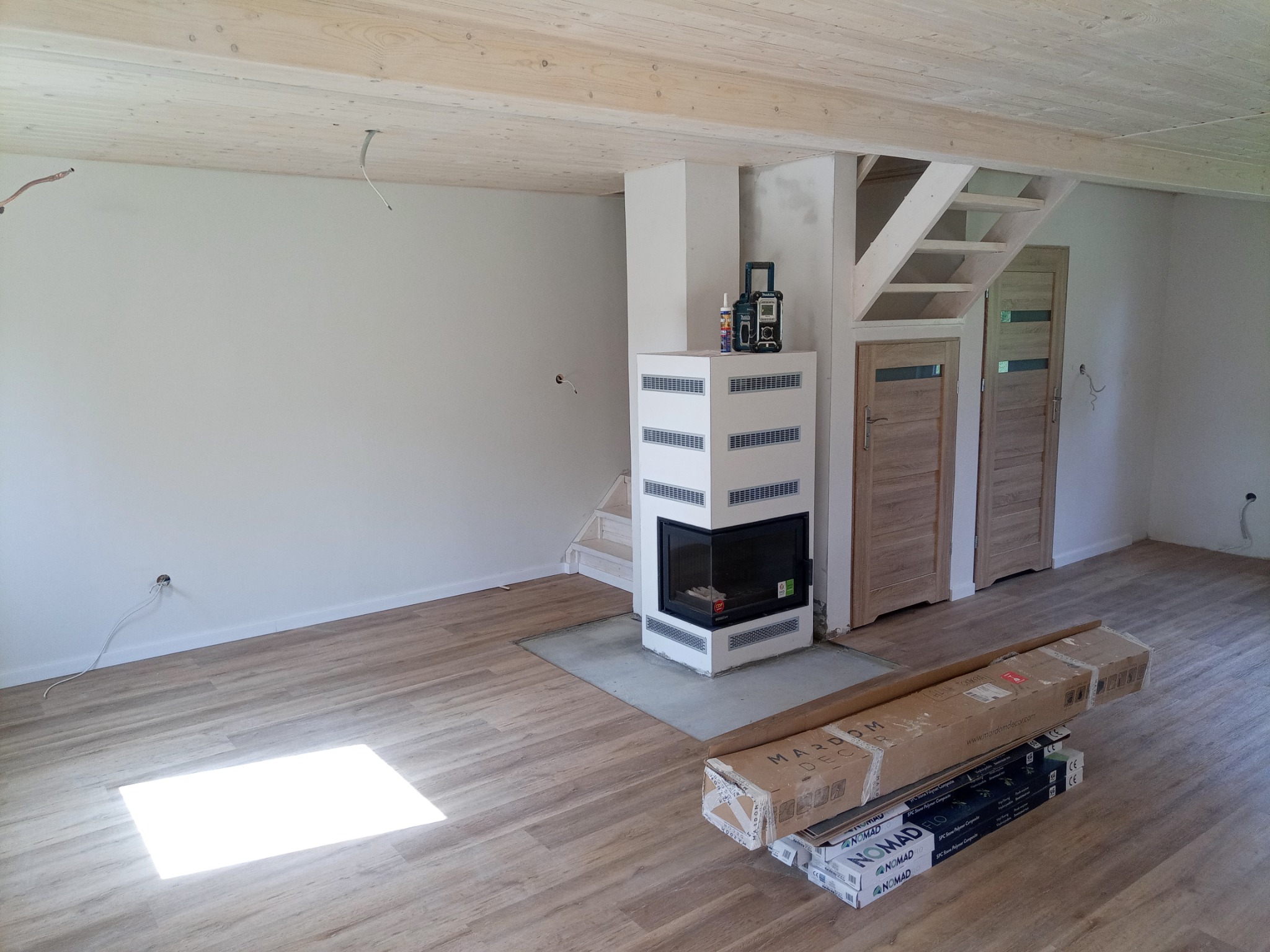 Wnętrze w trakcie wykańczania z drewnianym sufitem, kominkiem obudowanym białymi płytami, drewnianymi schodami i dwiema parami drzwi, na podłodze panele i kartony z nowymi panelami.