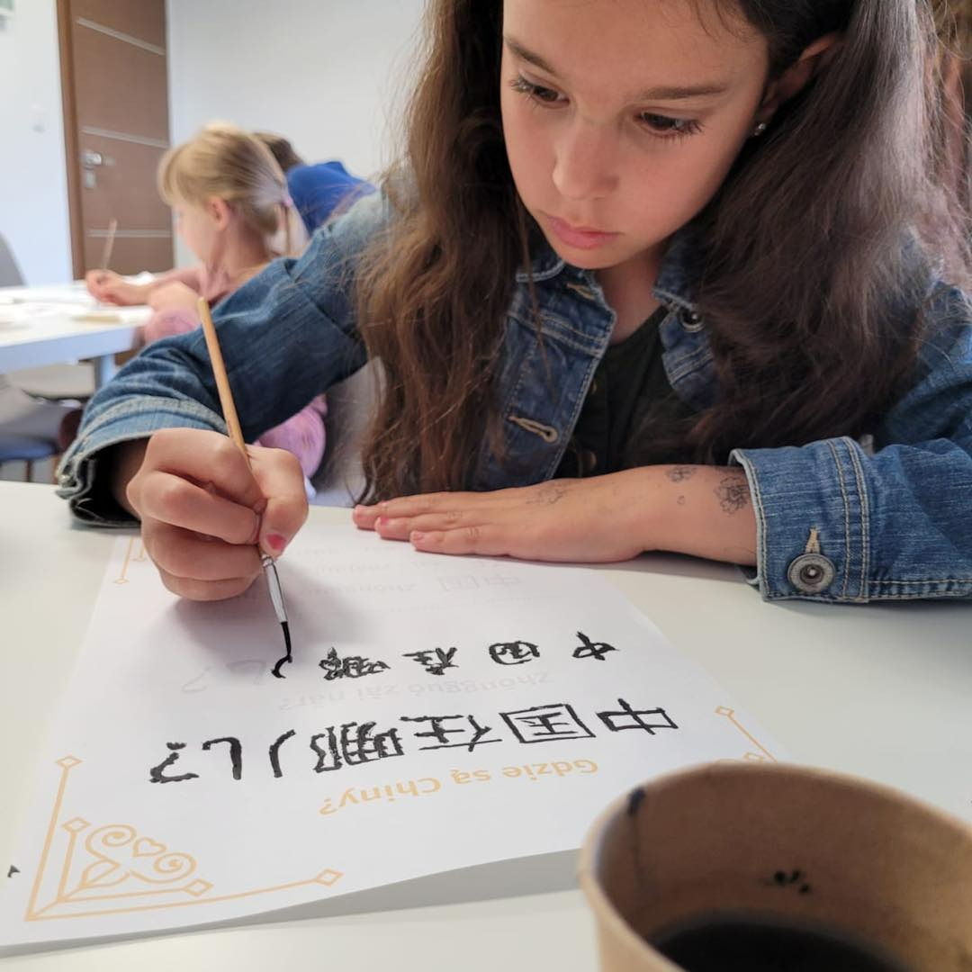 Dziewczynka w dżinsowej kurtce pisze pędzelkiem chińskie znaki na kartce z pytaniem 'Gdzie są Chiny?'