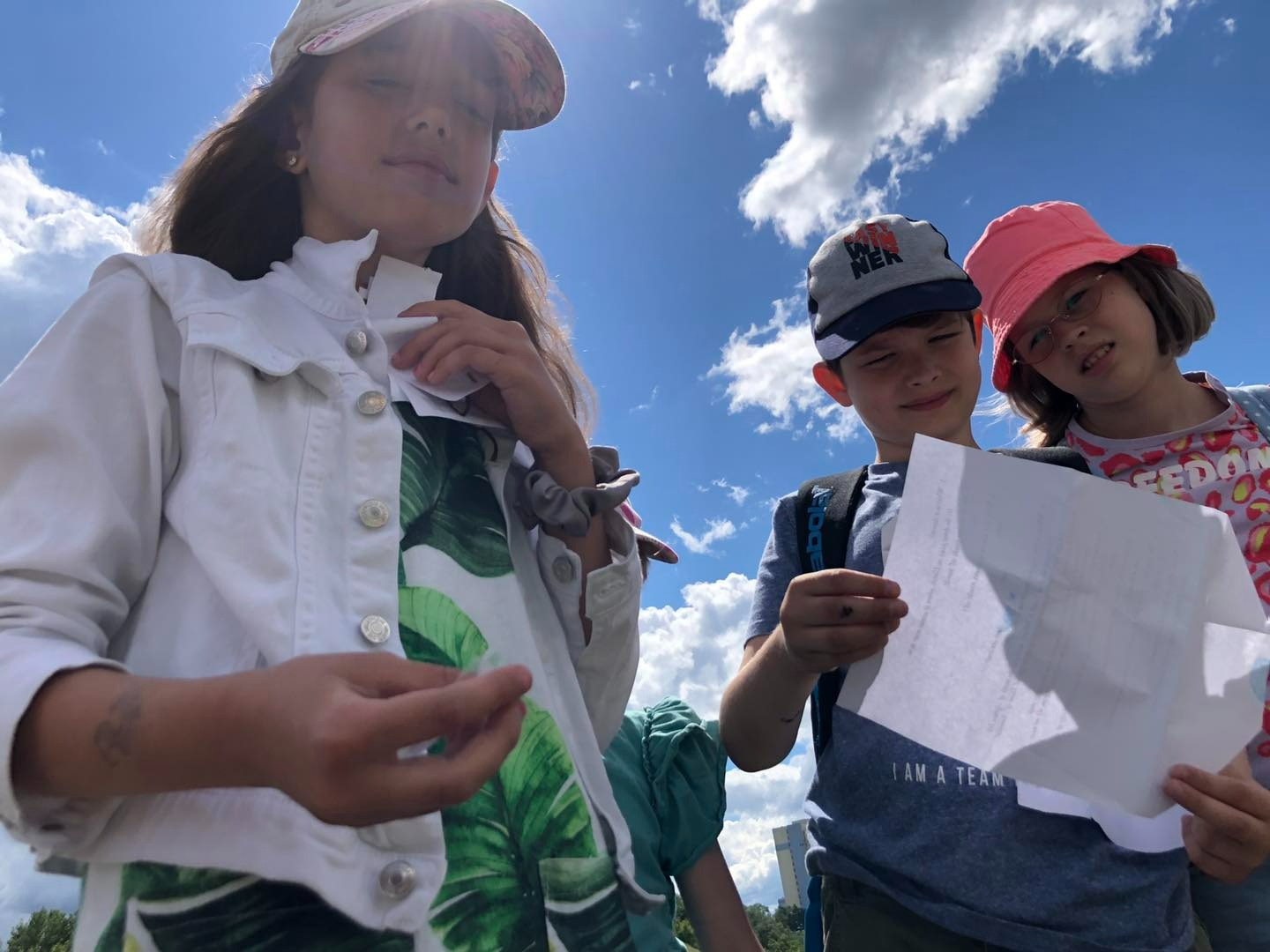 Troje dzieci spogląda w dół, trzymając kartkę z tekstem na tle błękitnego nieba z chmurami, prawdopodobnie podczas zajęć językowych na świeżym powietrzu.