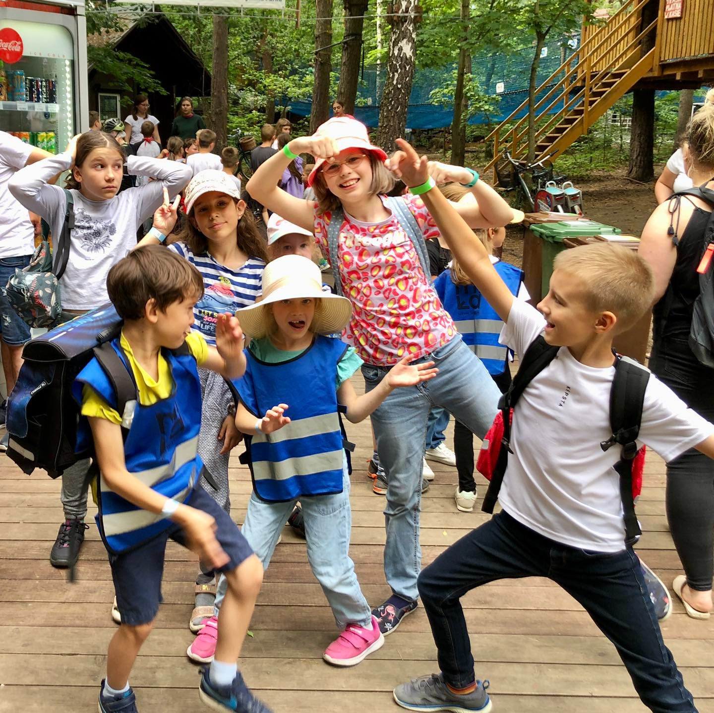 Grupa roześmianych dzieci w różnym wieku, w tym chłopcy i dziewczynki, pozuje na drewnianym podeście w otoczeniu drzew. Niektóre dzieci mają na sobie kamizelki odblaskowe i plecaki, a w tle...
