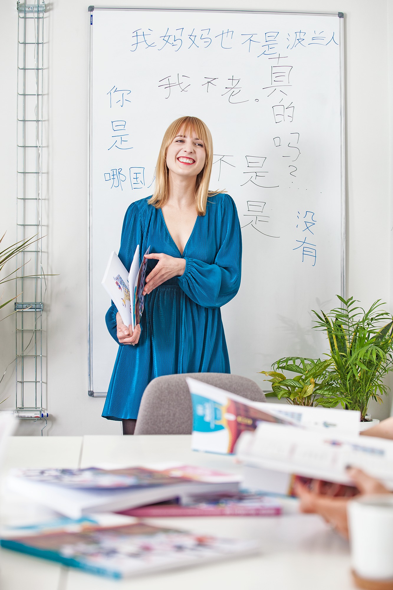 Uśmiechnięta nauczycielka w niebieskiej sukience trzymająca otwarty podręcznik na tle białej tablicy z napisanymi chińskimi znakami w klasie.