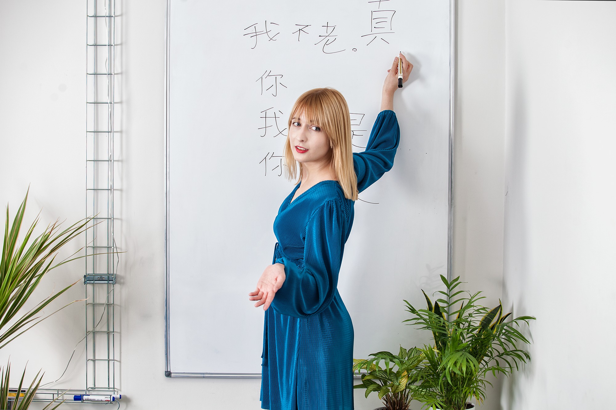 Nauczycielka z blond grzywką w niebieskiej sukience pisze markerem na białej tablicy wyrazy w języku chińskim, spoglądając w stronę widza.