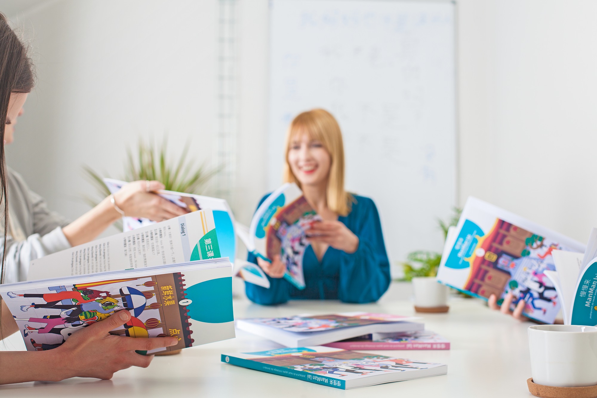 Nauczycielka z blond włosami uśmiecha się, trzymając otwarty podręcznik do nauki języka obcego, wokół leżą inne podręczniki i uczniowie przeglądają materiały.