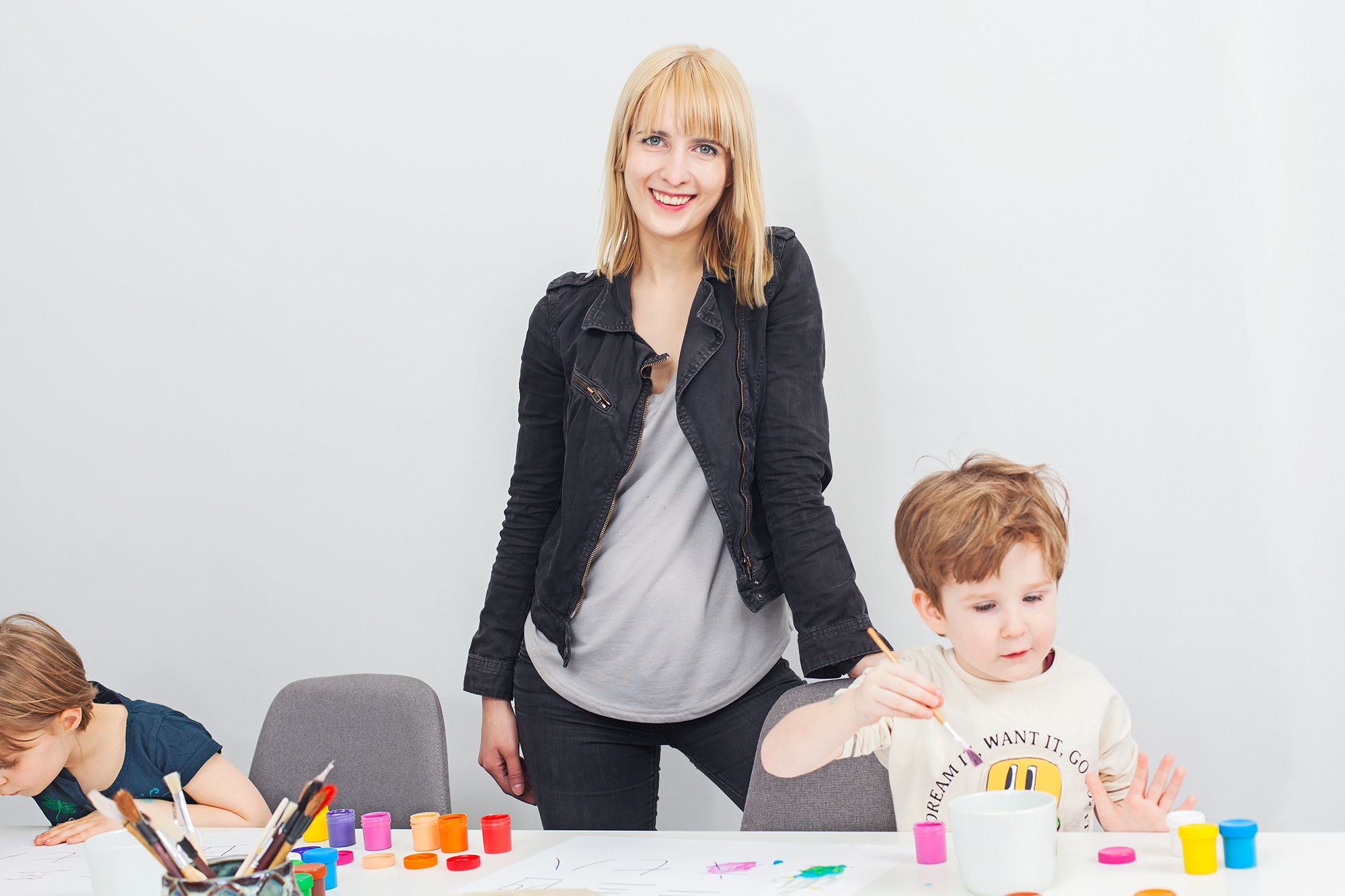 Uśmiechnięta blondynka w czarnej kurtce obserwuje dwóch chłopców malujących farbami na białym stole, jeden z nich trzyma pędzel w dłoni.