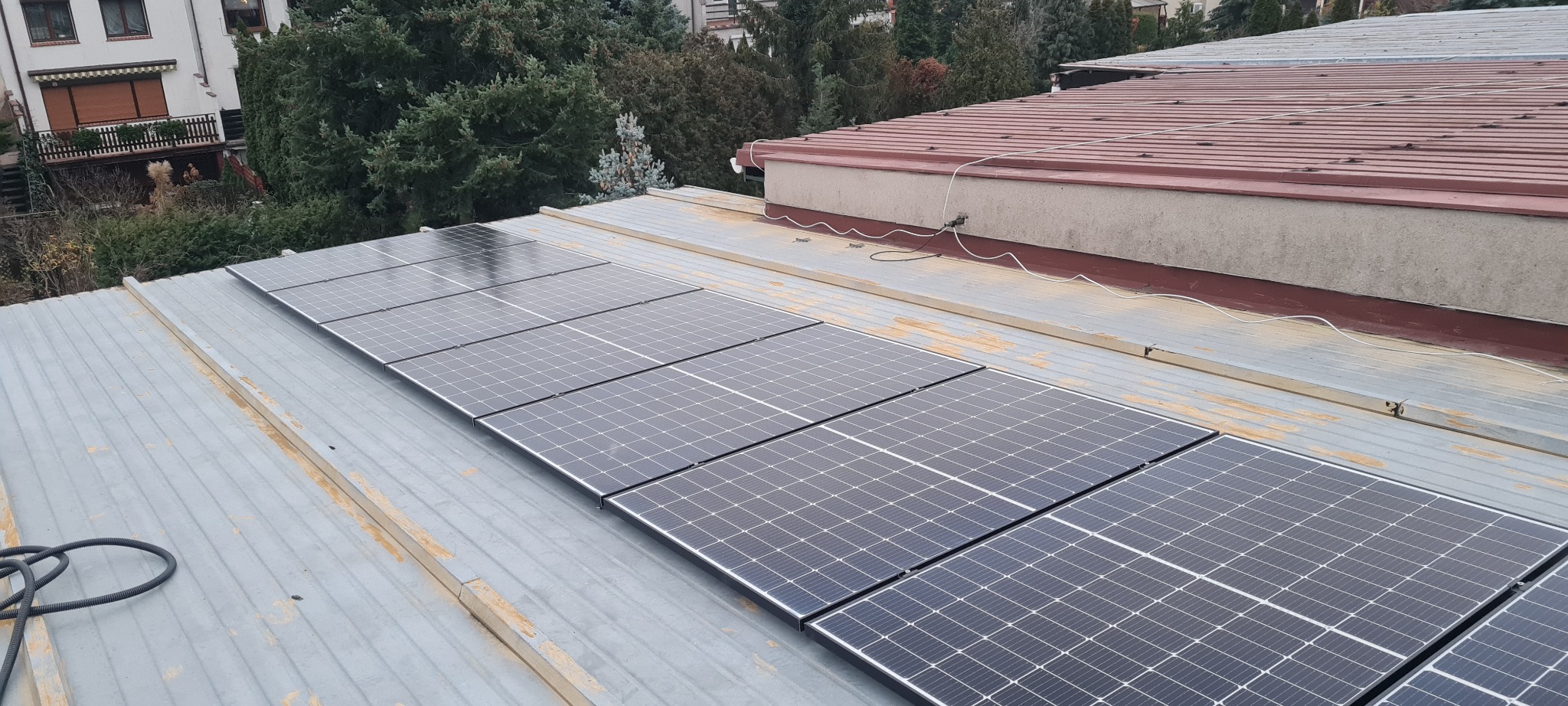 Panele słoneczne zamontowane na blaszanym dachu budynku mieszkalnego, widoczne przewody elektryczne i otoczenie z drzewami.
