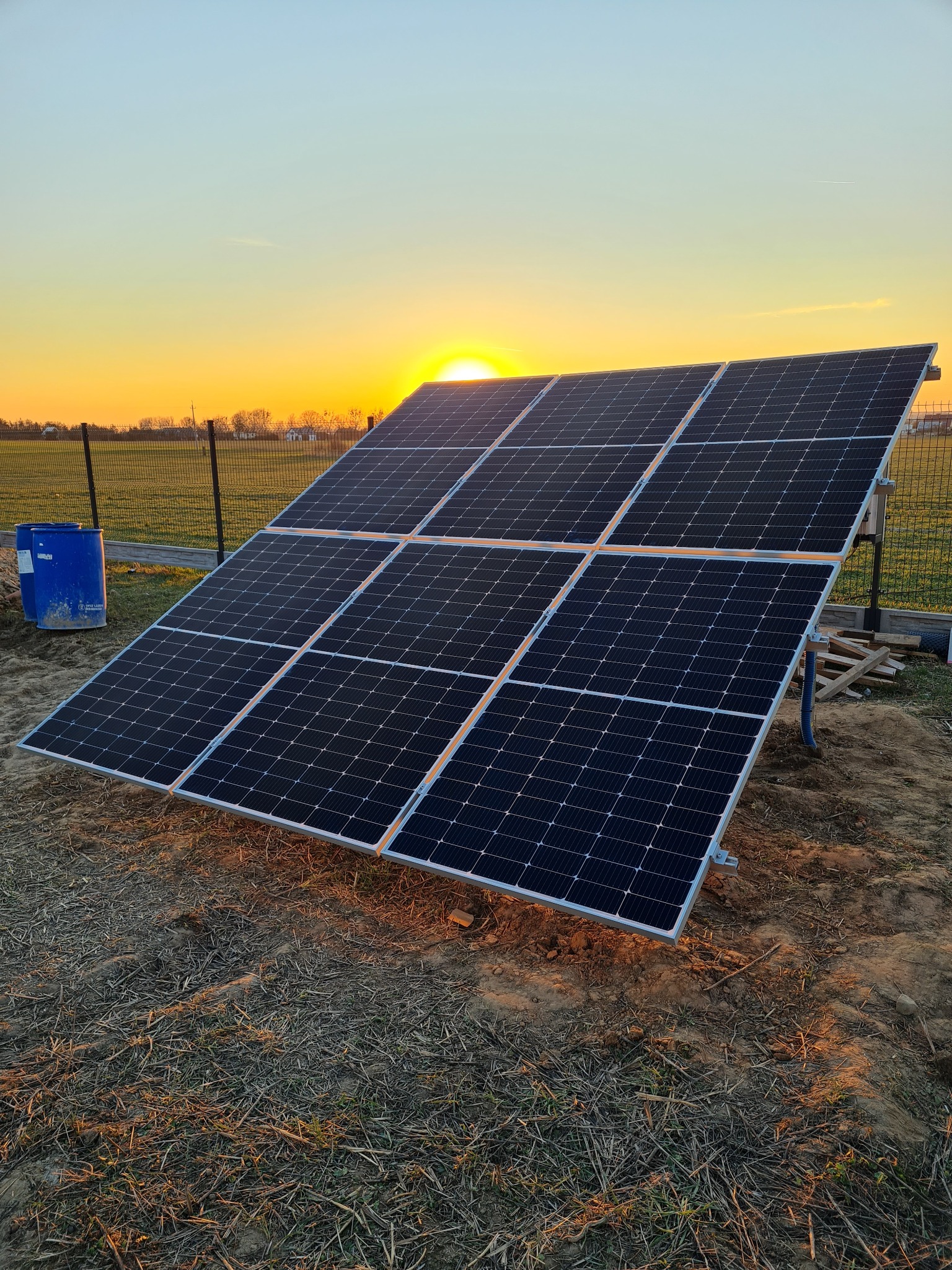 Panel fotowoltaiczny na polu, w tle zachodzące słońce i ogrodzenie z siatki, obok niebieskie beczki.