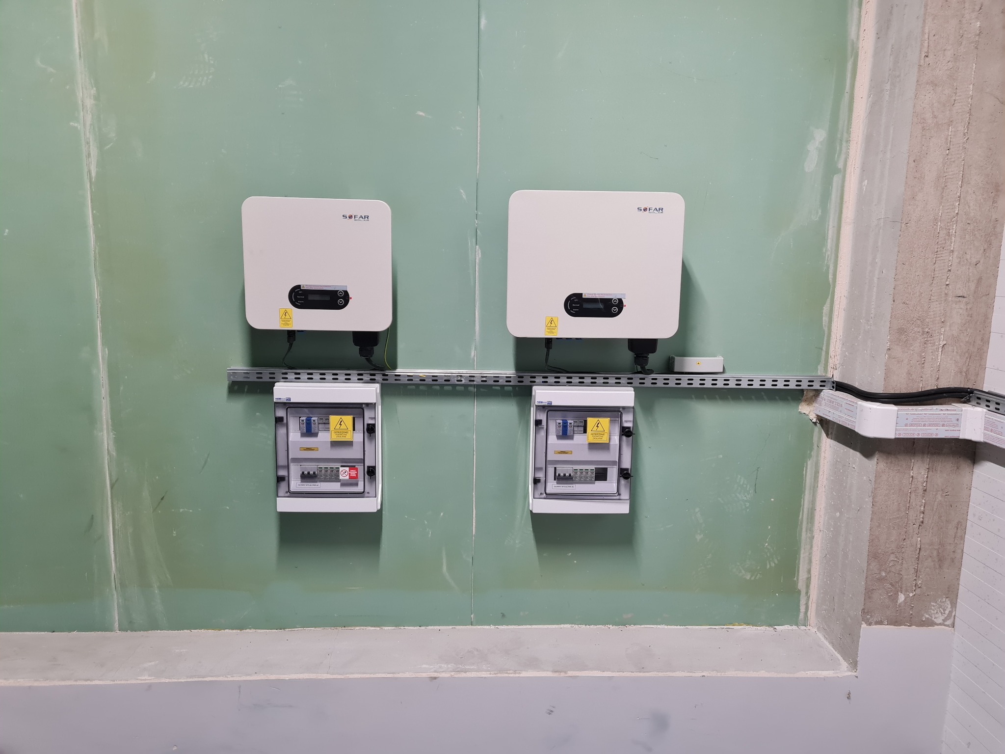 Dwa białe inwertery fotowoltaiczne marki SOFAR SOLAR zamontowane nad dwiema skrzynkami elektrycznymi na zielonej ścianie w Luboniu, widoczne okablowanie i elementy instalacji.