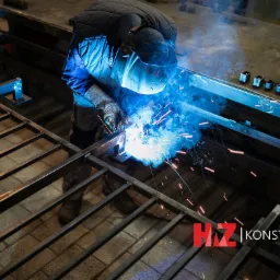 Spawanie metalowej konstrukcji kratowej, widoczne iskry i ochronna maska spawacza, logo firmy w prawym dolnym rogu.
