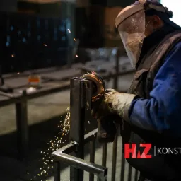 Pracownik w ochronnej masce szlifuje metalową konstrukcję, iskry rozbryzgują się w warsztacie.