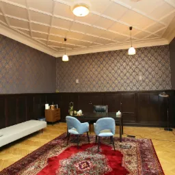 Wnętrze gabinetu z drewnianą boazerią, wzorzystą tapetą, dywanem orientalnym i dwoma błękitnymi krzesłami przy ciemnym biurku, uzupełnione kozetką i lampami wiszącymi.