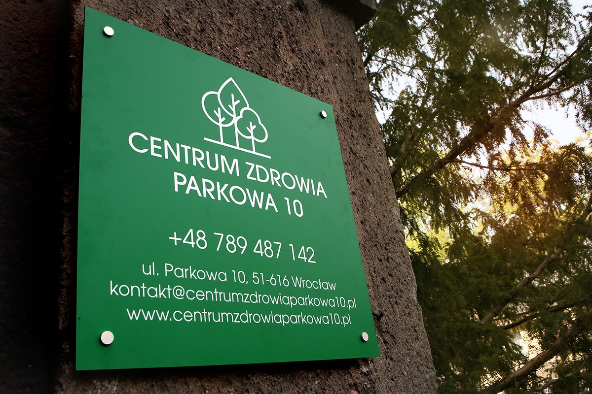 Zielona tablica informacyjna Centrum Zdrowia Parkowa 10 z logo drzew, numerem telefonu, adresem i adresem strony internetowej, przymocowana do pnia drzewa.