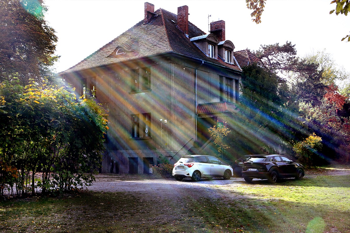 Dwupiętrowy budynek z czerwonym dachem w jesiennym słońcu, z dwoma samochodami zaparkowanymi przed budynkiem.