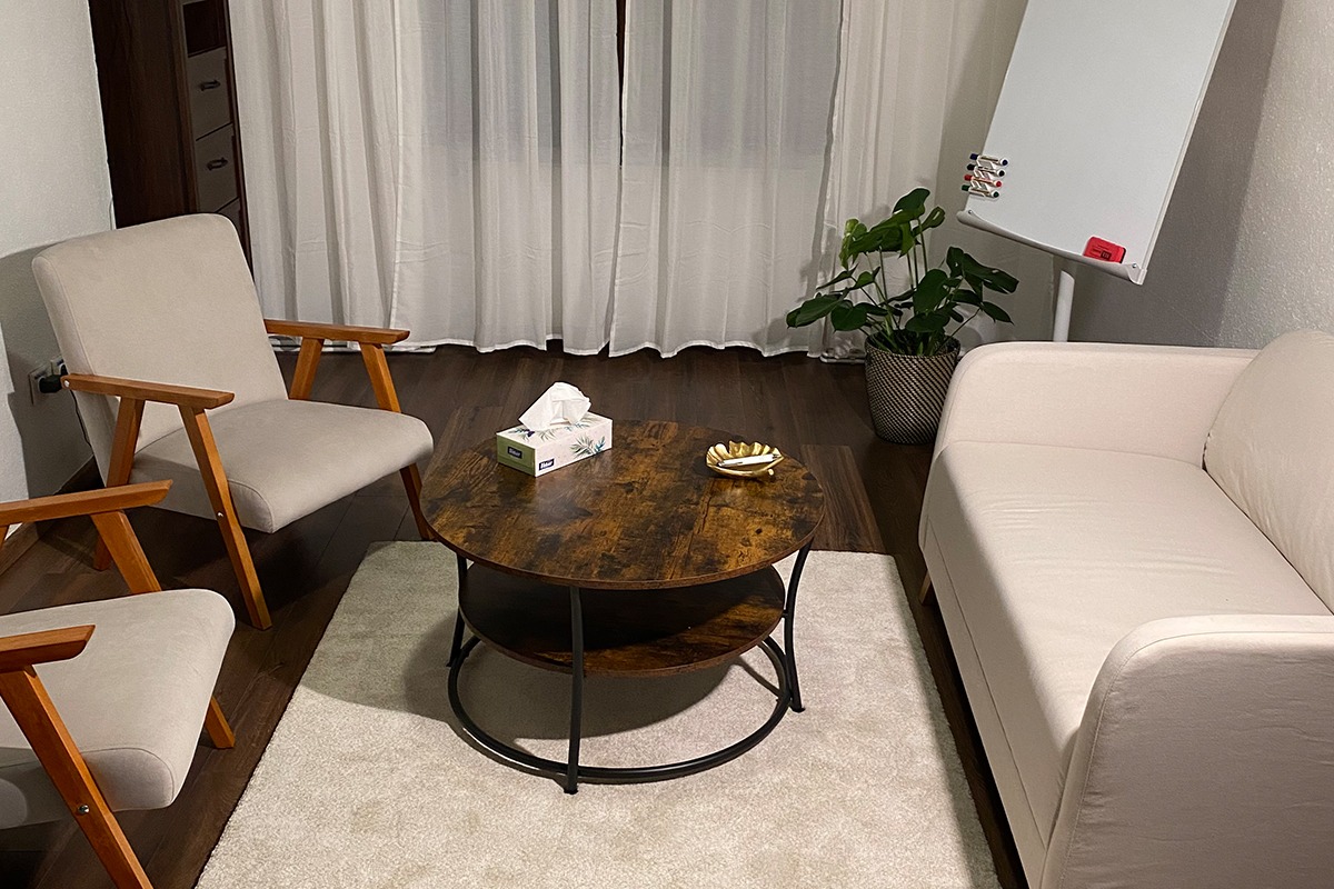 Jasne, minimalistyczne wnętrze gabinetu z dwoma fotelami z drewnianymi podłokietnikami, okrągłym stolikiem z chusteczkami i popielniczką, białą sofą oraz tablicą flipchart z markerami, oświetlone...