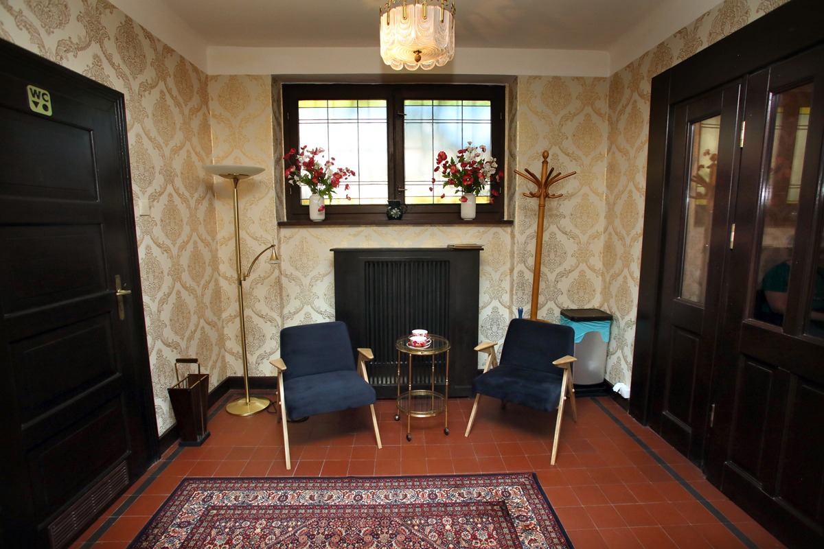 Poczekalnia z dwoma granatowymi fotelami, stolikiem z filiżankami, wzorzystą tapetą i dywanem w orientalnym stylu.