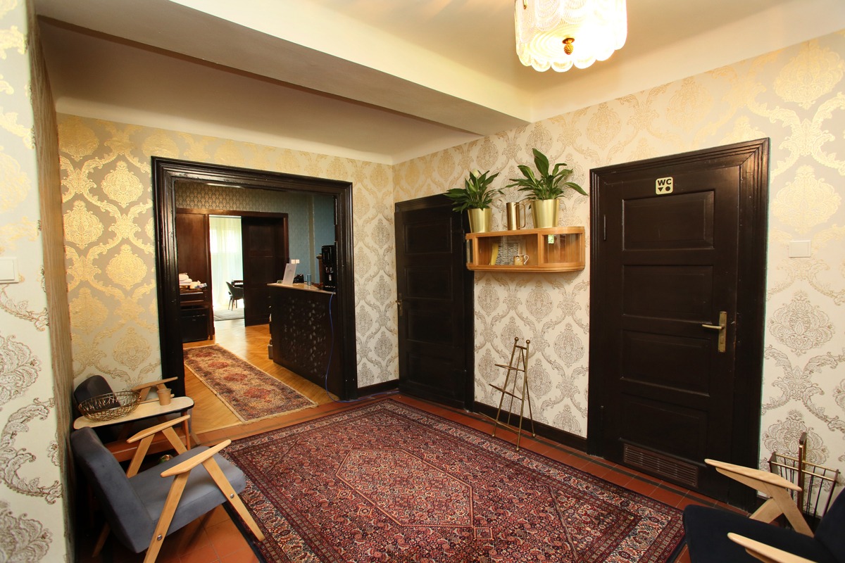 Wnętrze poczekalni: wzorzysta tapeta, dywan orientalny, krzesła w stylu retro, rośliny doniczkowe w złotych osłonkach, drzwi z oznaczeniem WC.