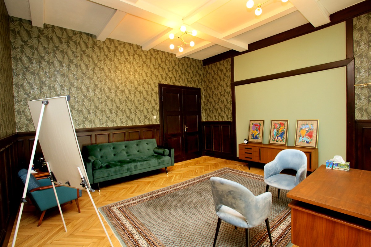 Gabinet z zieloną sofą, dwoma błękitnymi fotelami, biurkiem, komodą z obrazami i flipchartem, utrzymany w stylu retro z wzorzystą tapetą.