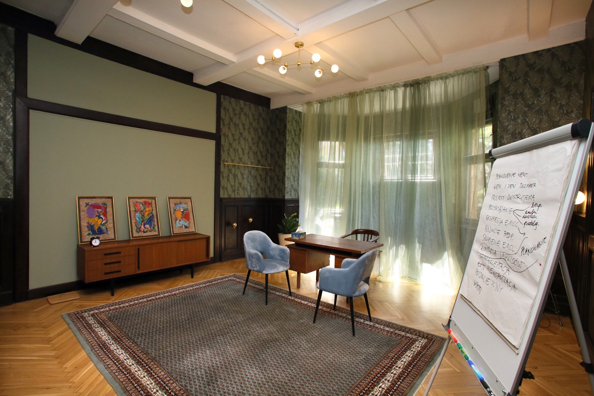 Wnętrze gabinetu z biurkiem, dwoma błękitnymi krzesłami, dywanem i flipchartem z zapiskami, utrzymane w stonowanej kolorystyce z elementami retro.