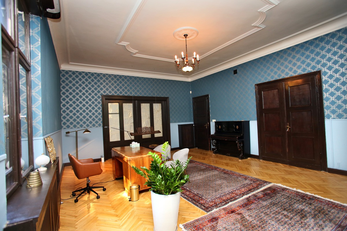 Gabinet z drewnianym biurkiem, brązowym krzesłem obrotowym, pianinem, dywanami orientalnymi i rośliną doniczkową na parkiecie w jodełkę, oświetlony żyrandolem.