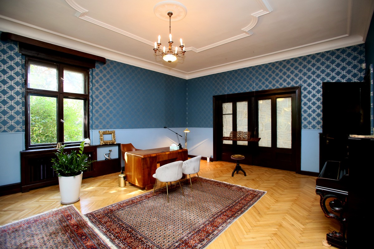 Gabinet z biurkiem, krzesłami, dywanem, pianinem i lampą, utrzymany w stylu retro z tapetami w geometryczny wzór.