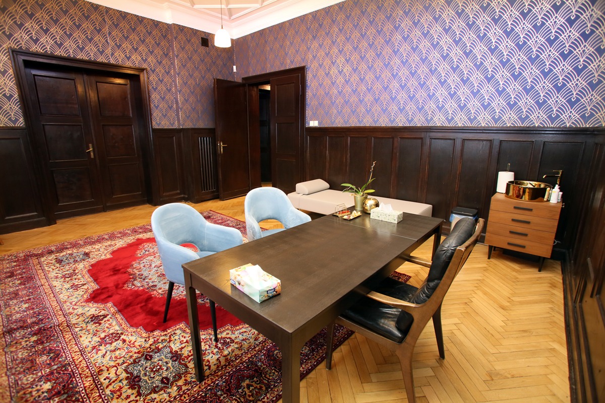 Wnętrze gabinetu z ciemnymi drewnianymi boazeriami, wzorzystą tapetą w odcieniach fioletu i złota, dywanem w orientalne wzory, drewnianym stołem, błękitnymi krzesłami i leżanką.
