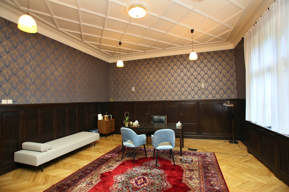 Wnętrze gabinetu z drewnianą boazerią, wzorzystą tapetą, dywanem orientalnym i dwoma błękitnymi krzesłami przy ciemnym biurku, uzupełnione kozetką i lampami wiszącymi.