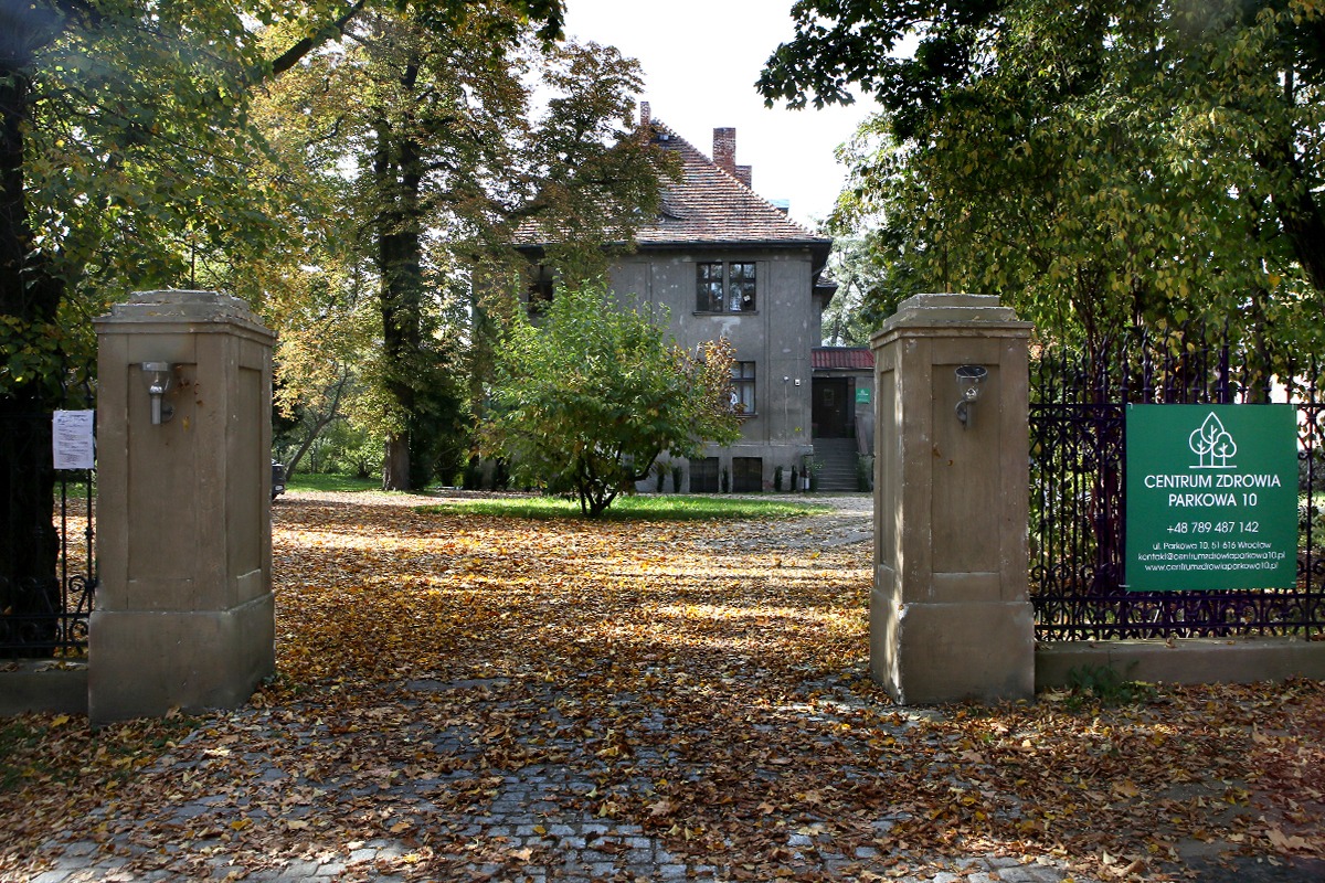 Wejście do Centrum Zdrowia Parkowa 10 w jesiennej scenerii, widoczne dwa kamienne słupki z kranami, żelazna brama i tablica informacyjna z logo, w tle budynek z czerwonym dachem.