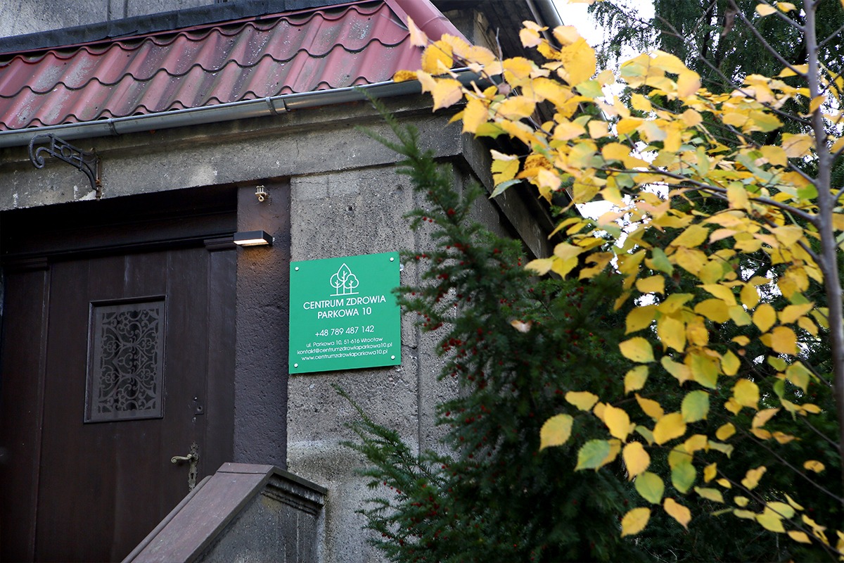 Wejście do Centrum Zdrowia Parkowa 10, widoczna tabliczka informacyjna z numerem telefonu i adresem strony internetowej, jesienne liście na drzewie, szare ściany budynku i brązowe drzwi z ozdobnym...
