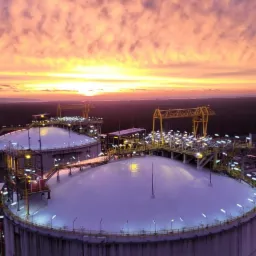 Dwa zbiorniki przemysłowe oświetlone w nocy, z pomarańczowym zachodem słońca w tle i zalesionym krajobrazem w oddali.