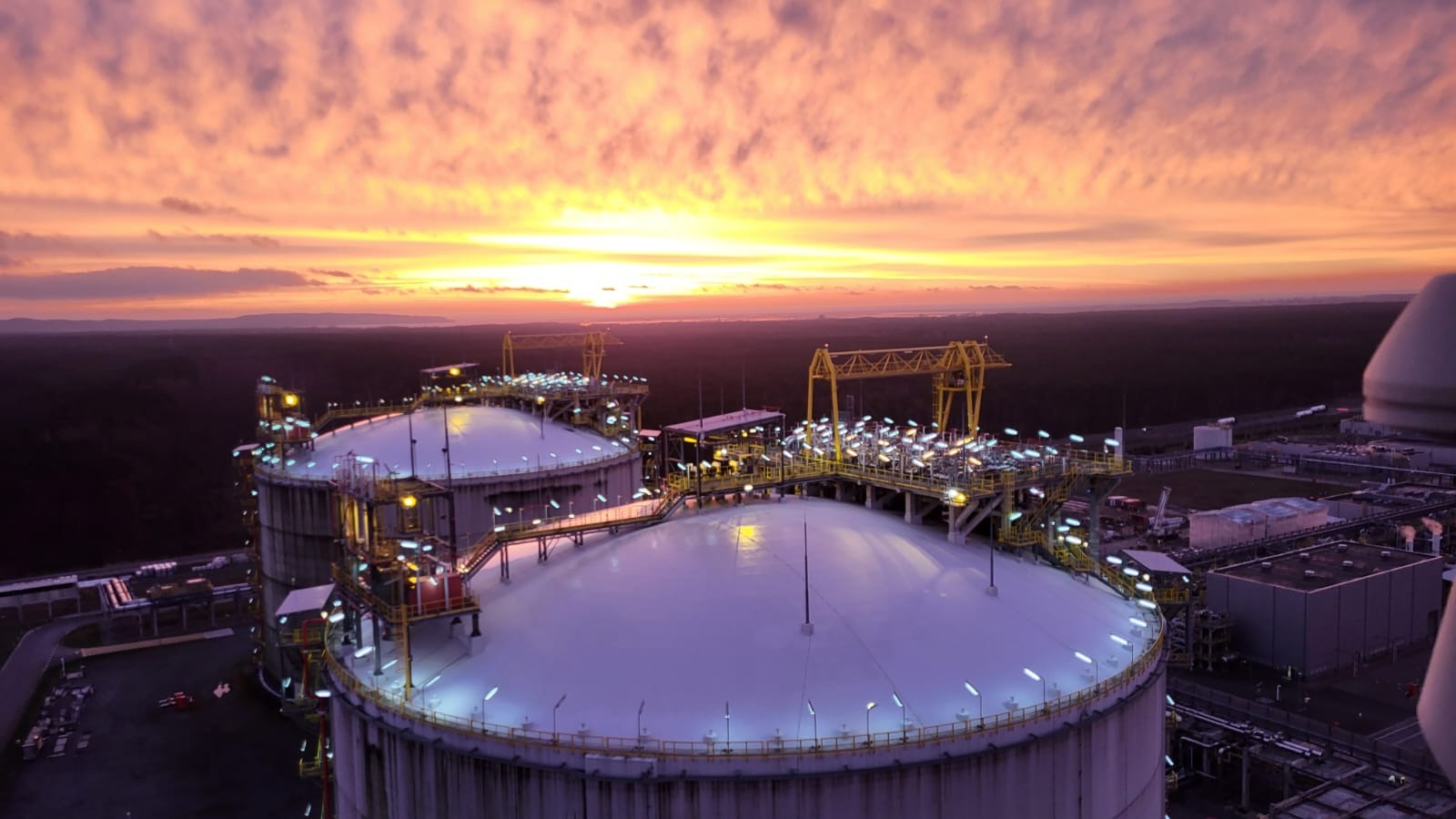 Dwa zbiorniki przemysłowe oświetlone w nocy, z pomarańczowym zachodem słońca w tle i zalesionym krajobrazem w oddali.