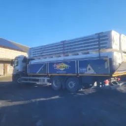 Ciężarówka z plandeką firmy Blachy Pruszyński przewożąca panele dachowe w opakowaniu, na tle budynku z panelami słonecznymi na dachu.