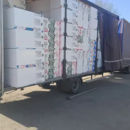 Ciężarówka wypełniona po brzegi białymi paczkami materiałów izolacyjnych z widocznymi etykietami producenta, częściowo zasłonięta plandeką, na nierównym terenie.