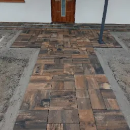 Ułożona ścieżka z brązowo-czarnej kostki przed wejściem do domu z brązowymi drzwiami, widoczne obrzeża i przygotowane podłoże.