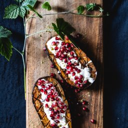 TLEN FOTO STUDIO - Krzysztof Tlen - Dwa przekrojone bakłażany ułożone na drewnianej desce, polane białym sosem, posypane pestkami granatu i ziołami, obok gałązka z zielonymi liśćmi.