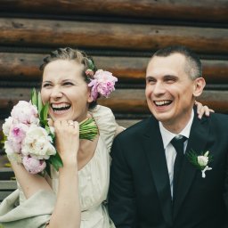 TLEN FOTO STUDIO - Krzysztof Tlen - Uśmiechnięta panna młoda w jasnej sukni trzyma bukiet piwonii, obok elegancko ubrany pan młody na tle drewnianej ściany.