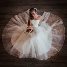 Panna młoda w białej sukni, widziana z góry, siedzi na drewnianej podłodze, trzyma bukiet różowych i białych kwiatów, uśmiecha się, ma na sobie srebrny naszyjnik i delikatną ozdobę we włosach.