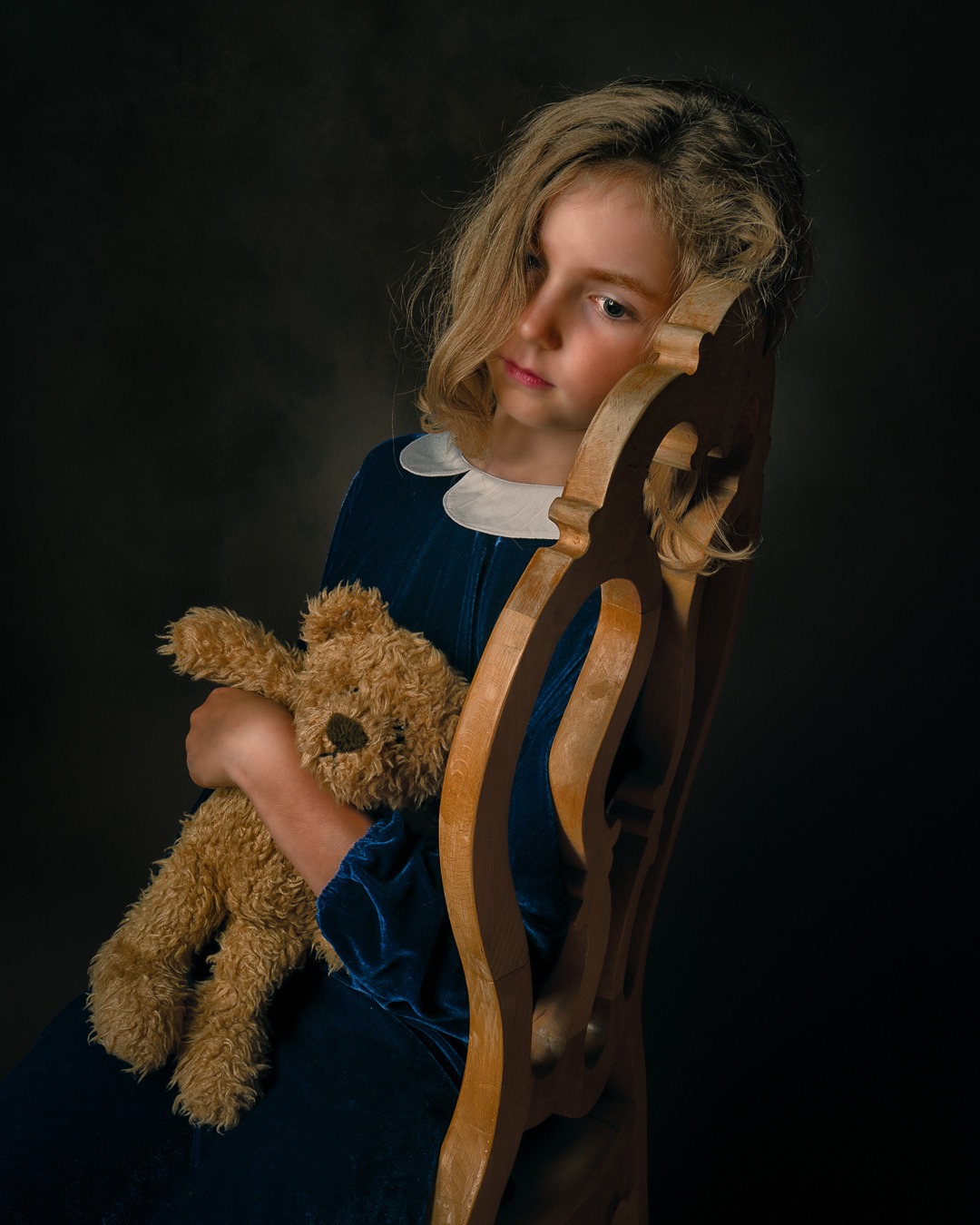 Portret dziewczynki w niebieskiej aksamitnej sukience z białym kołnierzykiem, siedzącej na drewnianym krześle i trzymającej pluszowego misia na ciemnym tle.
