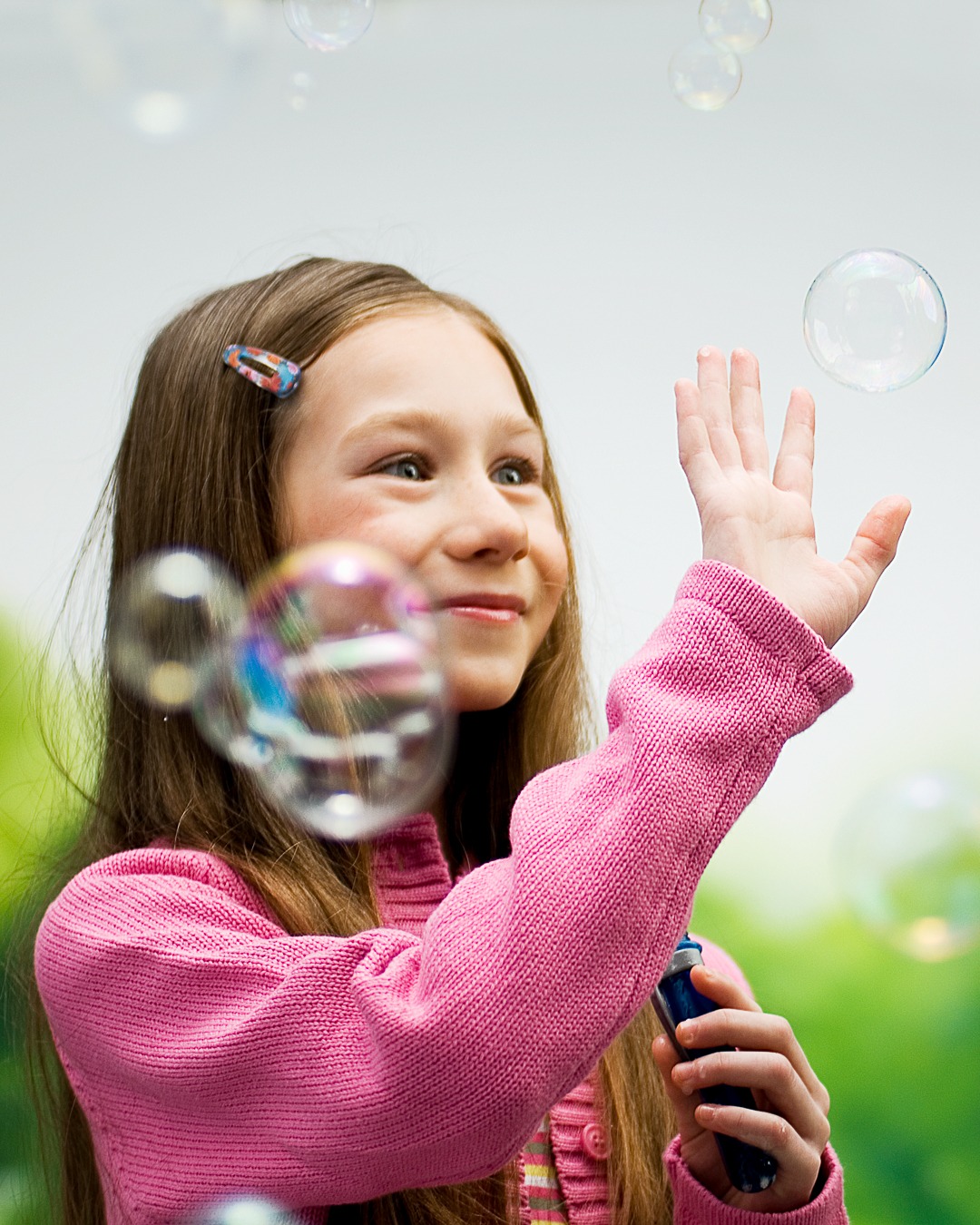 Uśmiechnięta dziewczynka w różowym swetrze puszcza bańki mydlane, trzymając w dłoni niebieski przyrząd do robienia baniek; w tle rozmyte zielone tło i unoszące się bańki.
