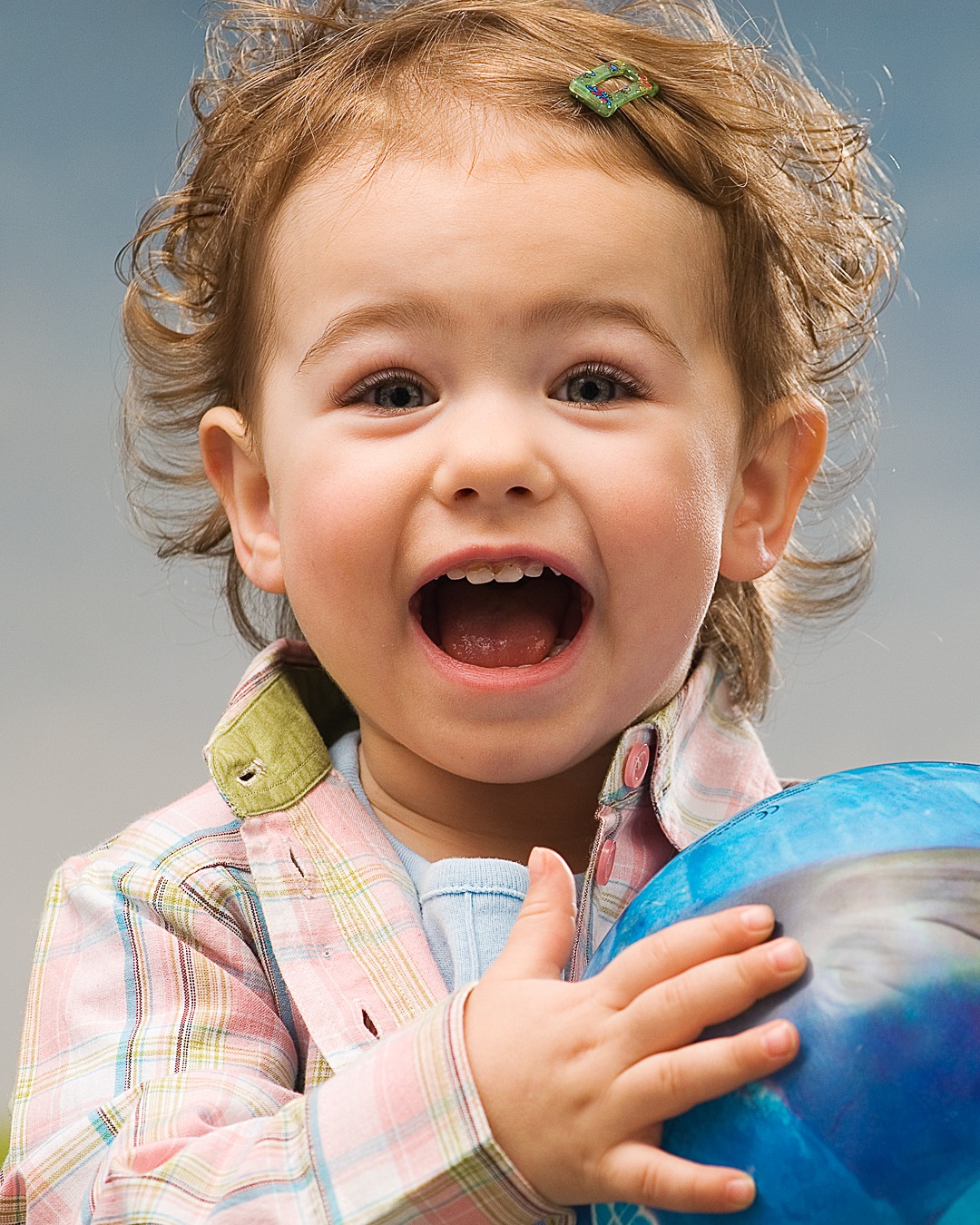 Uśmiechnięta dziewczynka z kręconymi włosami i spinką we włosach, trzymająca niebieską piłkę na rozmytym tle.