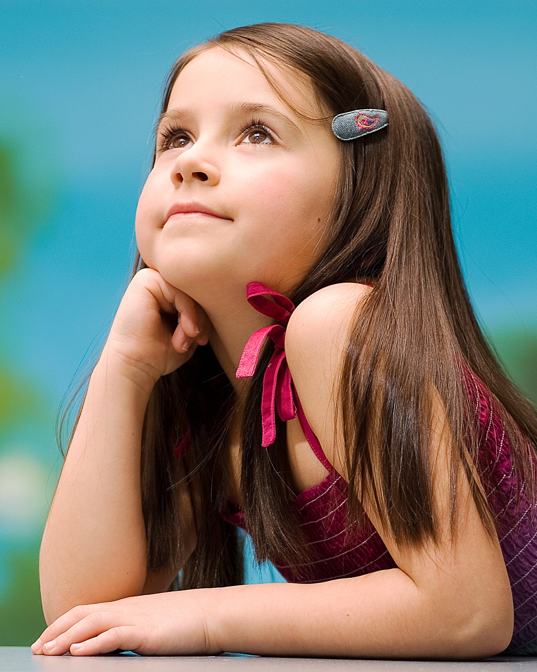 Portret młodej dziewczynki z brązowymi włosami, spoglądającej w górę, z ręką podpartą pod brodą, na tle błękitnego tła z delikatnym gradientem.