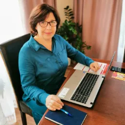 Kobieta w okularach siedzi przy drewnianym biurku z laptopem, notatnikiem i długopisem, uśmiechając się do kamery; w tle zasłony i roślina doniczkowa.