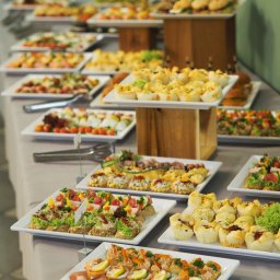 Cynamon Holding Sp. z o.o. - Długi stół nakryty szarym obrusem, zastawiony białymi kwadratowymi talerzami z różnorodnymi przekąskami: mini kanapeczki, tartaletki, roladki z szynki parmeńskiej i melona, ustawione na różnych...