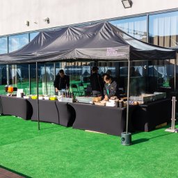 Cynamon Holding Sp. z o.o. - Zewnętrzny punkt gastronomiczny pod czarnym namiotem z widocznym logo firmy, obsługa w strojach roboczych przygotowuje dania na grillu, na stole pojemniki z jedzeniem i napojami, zielona sztuczna...